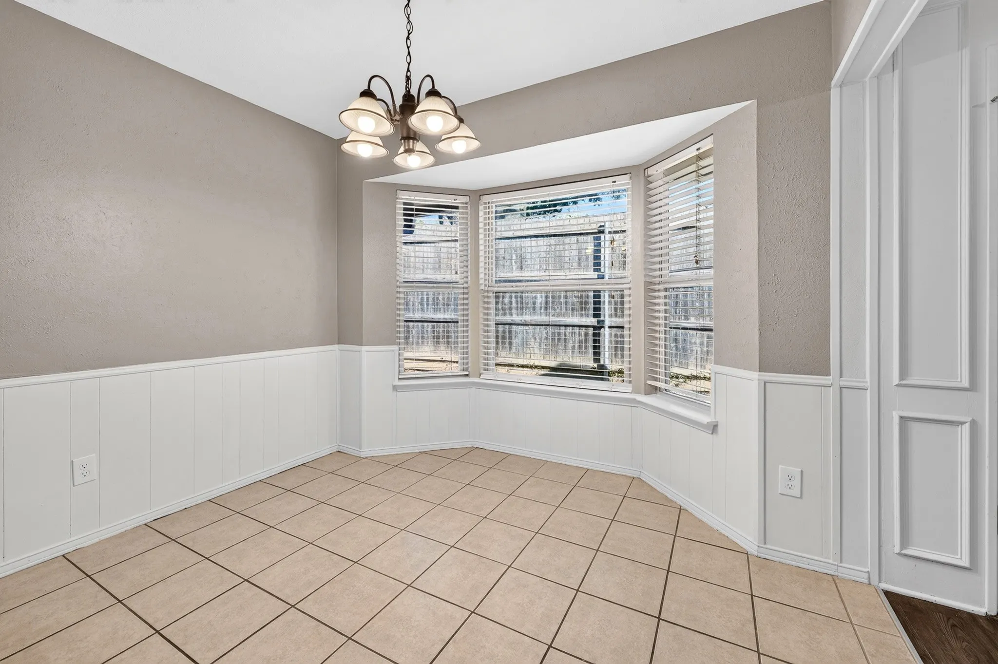 Bay window with slatted blinds, white wainscoting, neutral wall paint, ceiling-mounted light fixture, and ceramic tile flooring