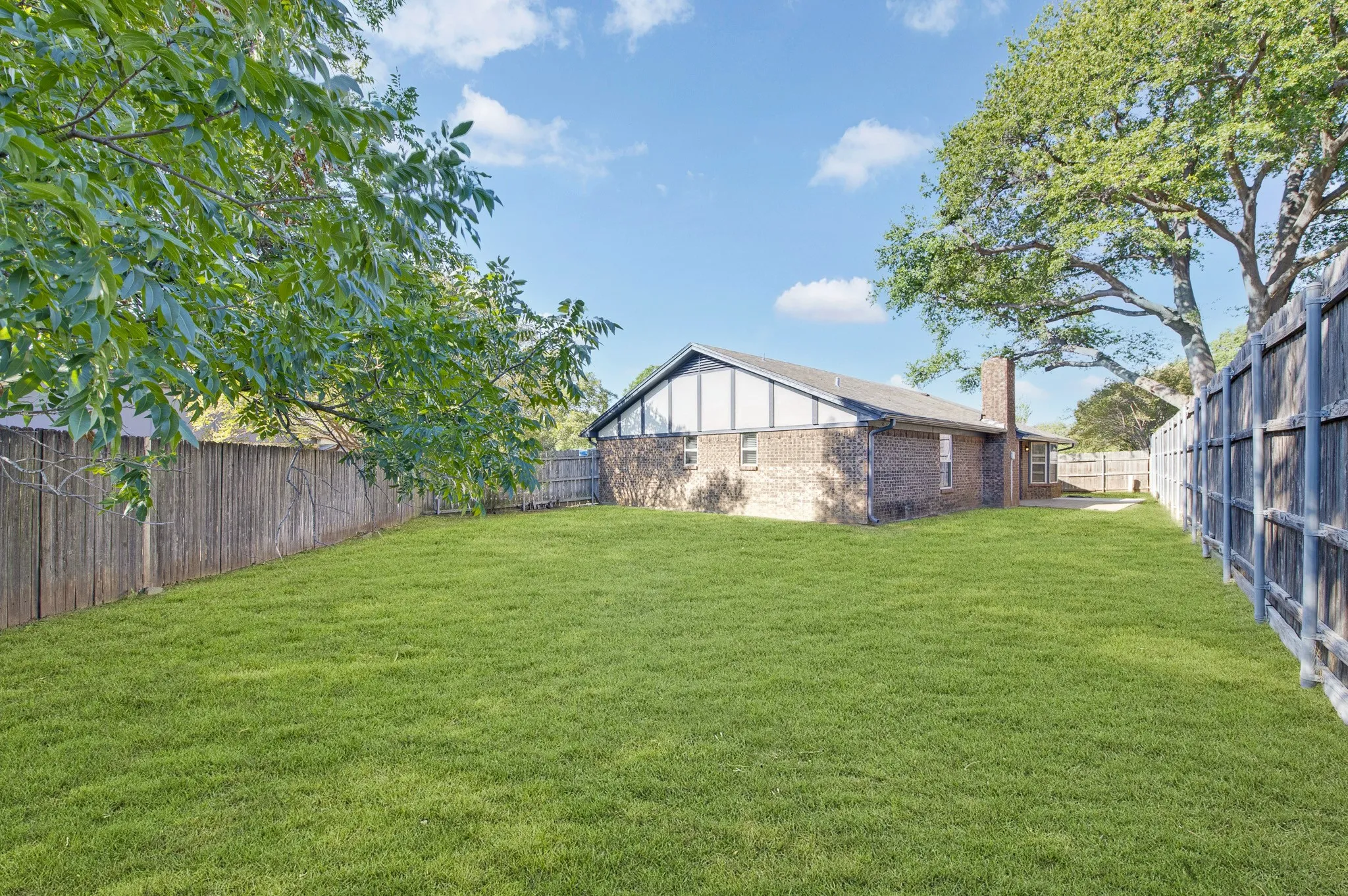 Expansive backyard featuring a manicured lawn, full perimeter fencing, mature trees, and a brick exterior with a pitched roof