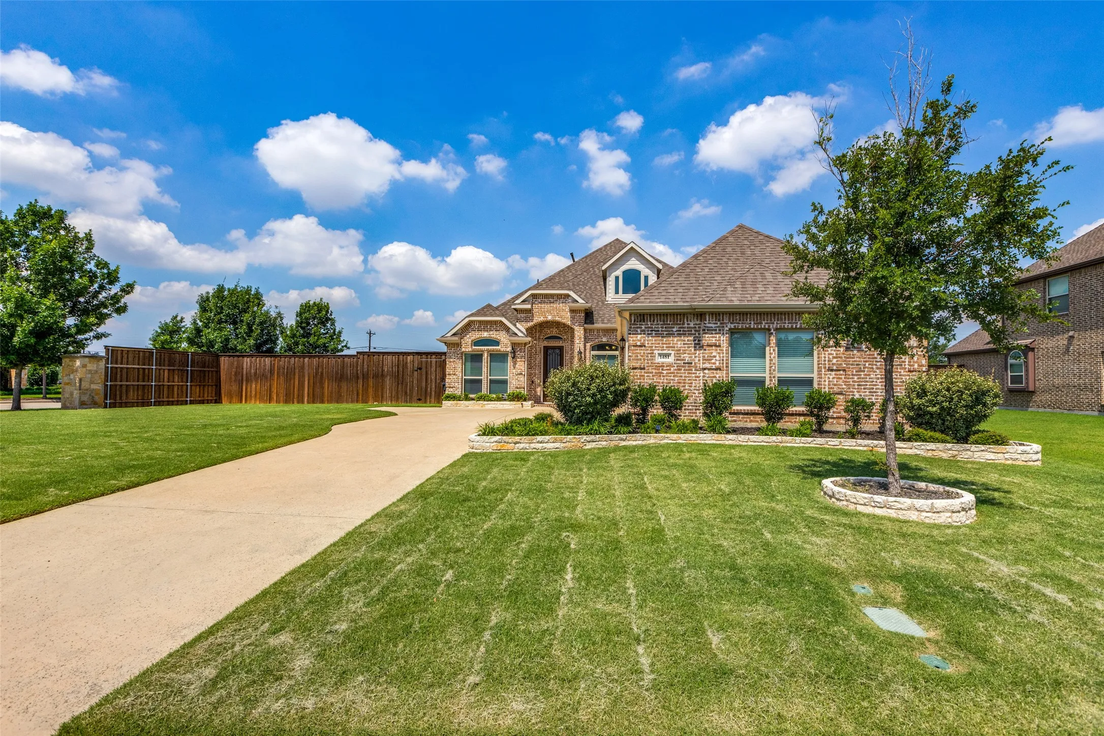 Brick exterior with stone accents, concrete driveway, front yard with green lawn, and wood privacy fence
