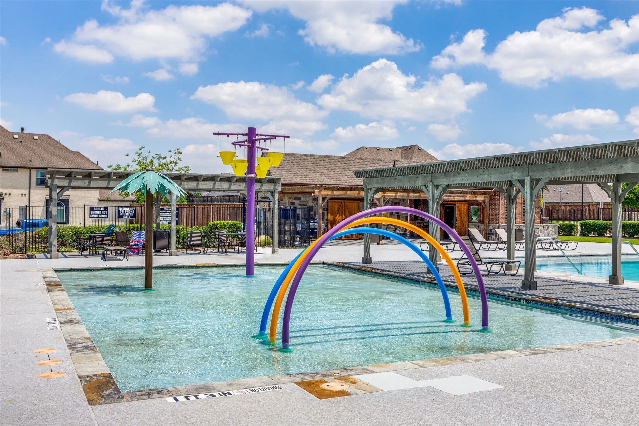 Community splash pad with multiple water features, adjacent to a swimming pool and covered pergola