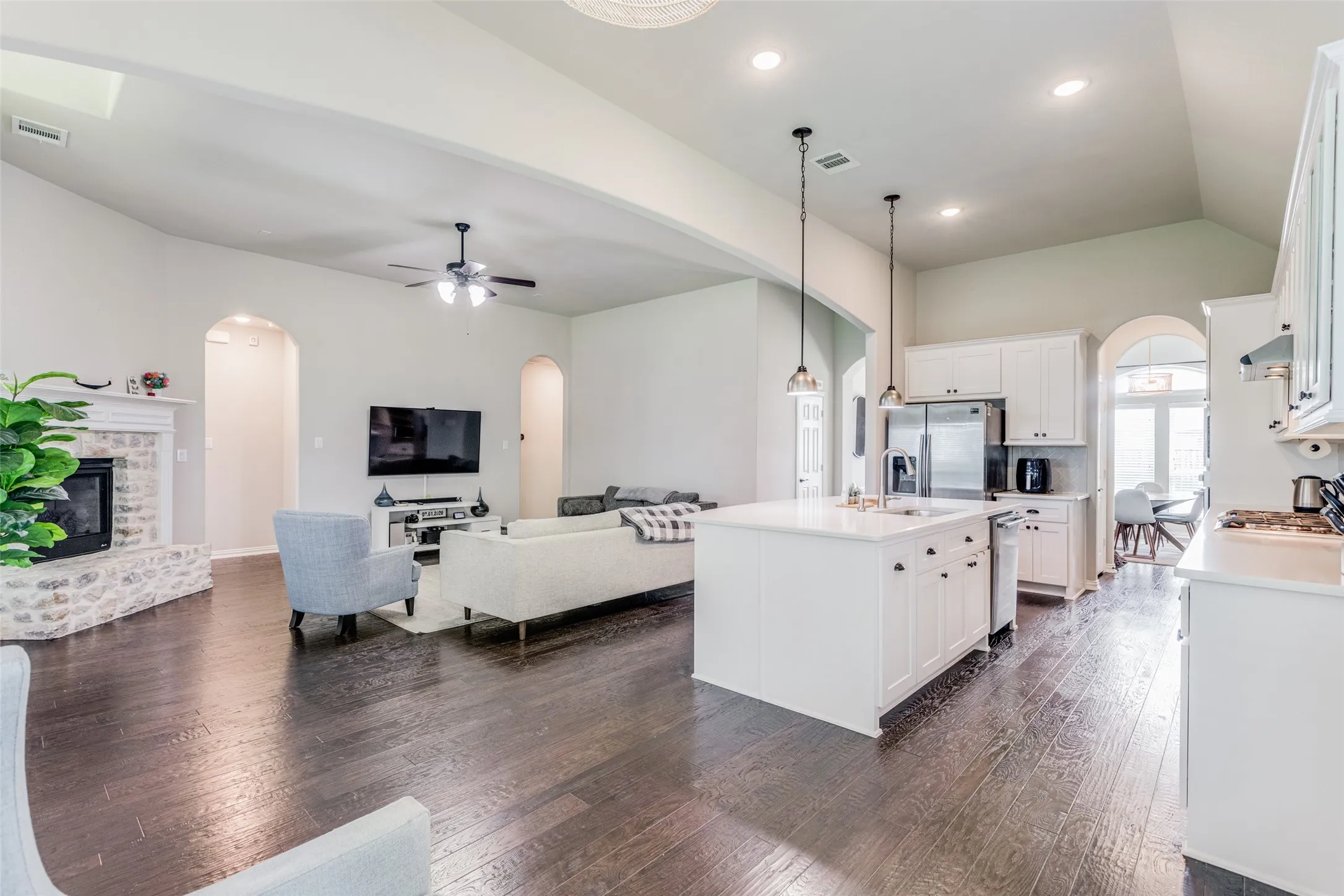 Open-concept living space featuring wood-finish flooring, a white kitchen island with sink, stainless steel appliances, a stone-clad fireplace, and arched doorways