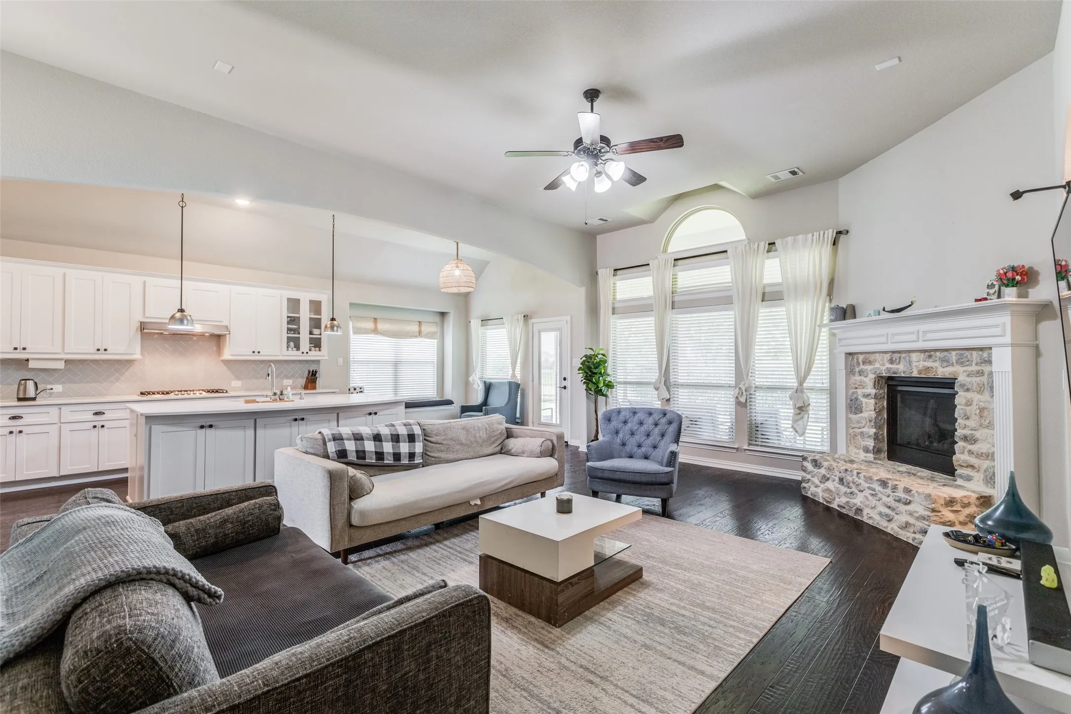 Wood-finish flooring, stone fireplace, vaulted ceiling with a fan, kitchen island with pendant lighting, and arched window