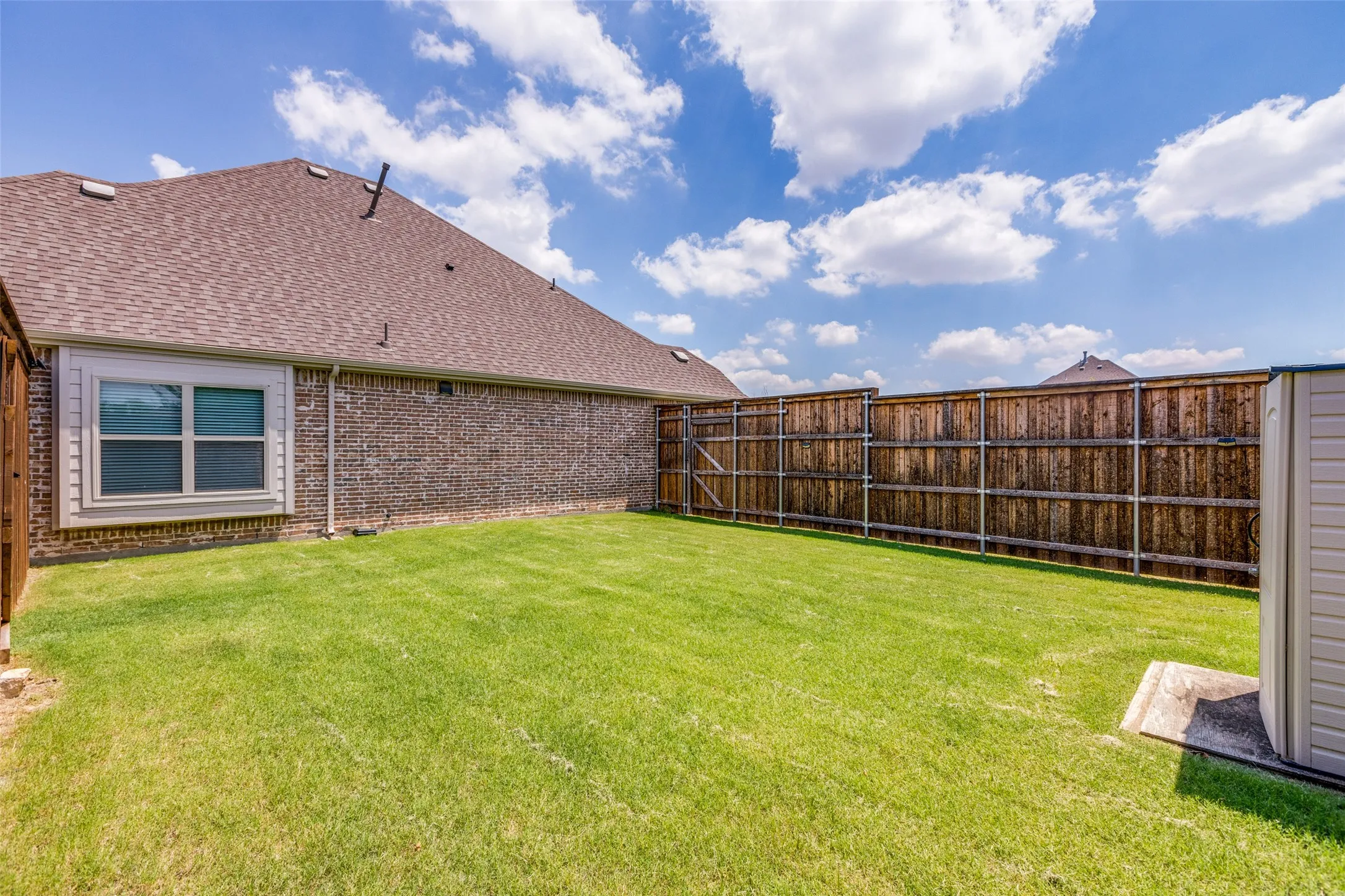Expansive grassy backyard featuring a full privacy fence, brick exterior, and a storage shed