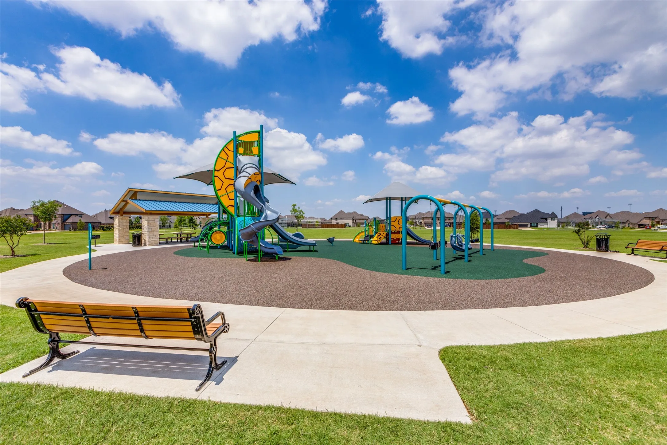 Community playground featuring multiple slides, swing sets, a covered picnic area, and a paved pathway
