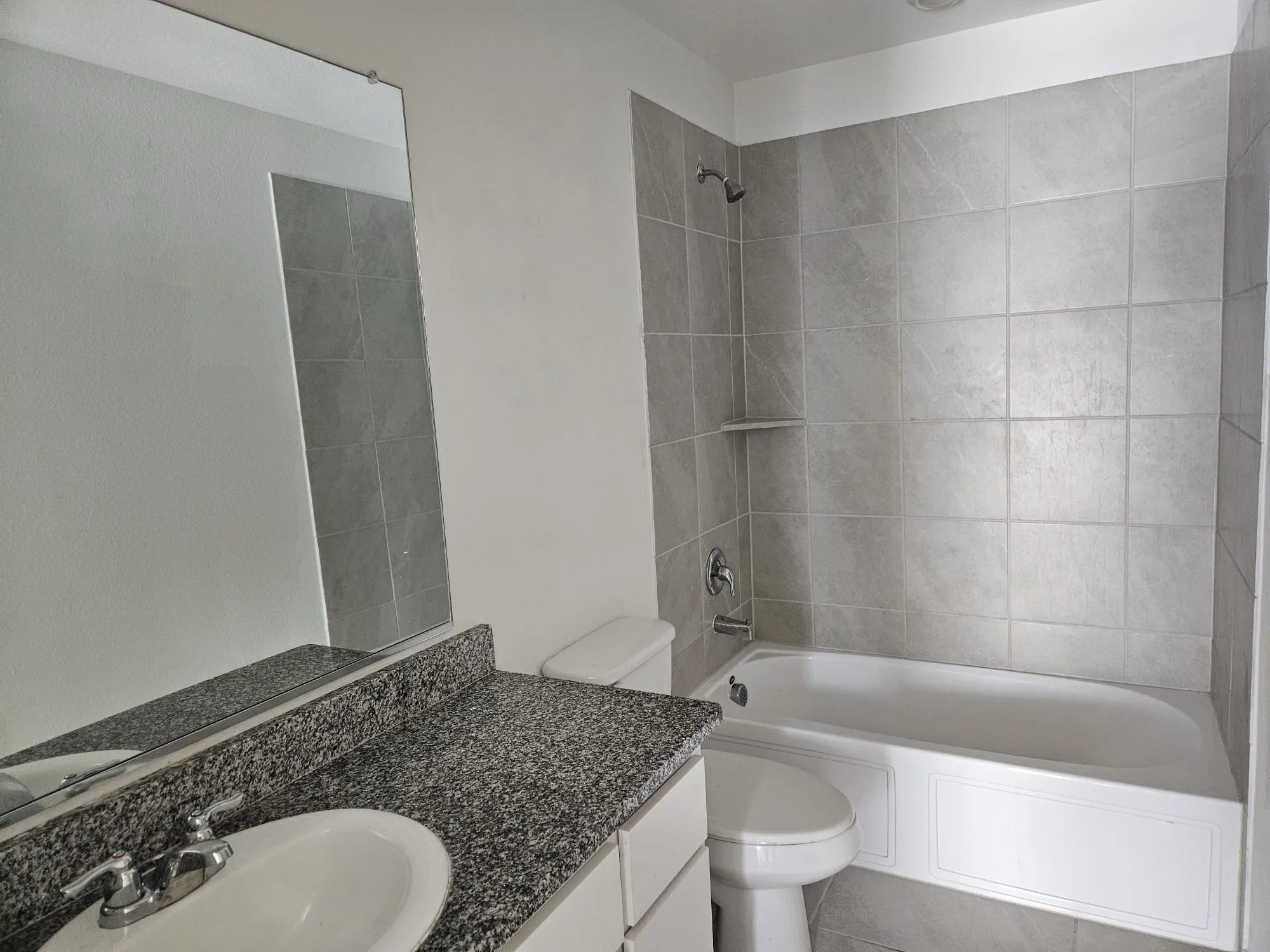 Bathroom featuring a white vanity with a speckled countertop, an integrated sink, a rectangular mirror, and a shower-over-tub with light gray tile surround