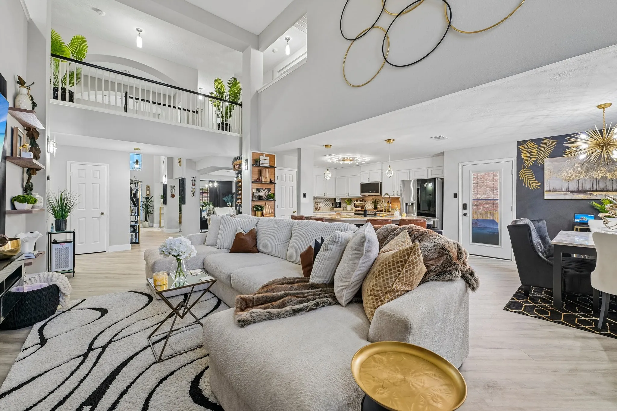 Open-concept living area featuring a double-height ceiling, upper-level balcony with white balustrade, wood-finish flooring, and a dining area with a gold chandelier