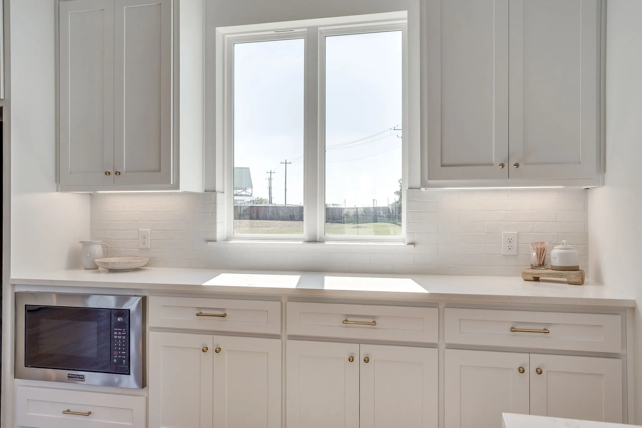 White cabinetry with gold-tone hardware, white countertops, built-in stainless steel microwave, and a white subway tile backsplash