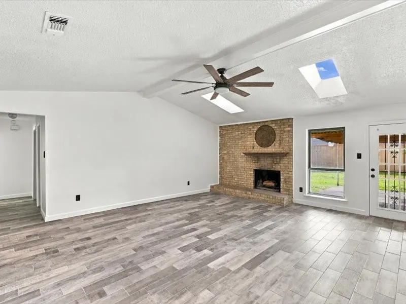 Vaulted ceiling featuring skylights and a ceiling fan, brick fireplace with a wood mantel, wood-finish flooring, and a glass-paned exterior door