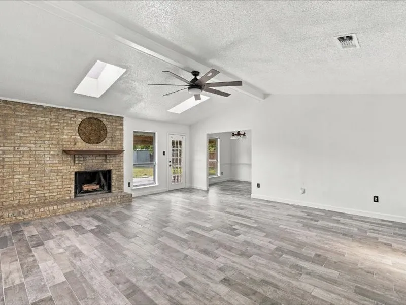 Vaulted ceiling with skylights, wood-finish flooring, brick fireplace with wood mantle, and a glass-paneled door