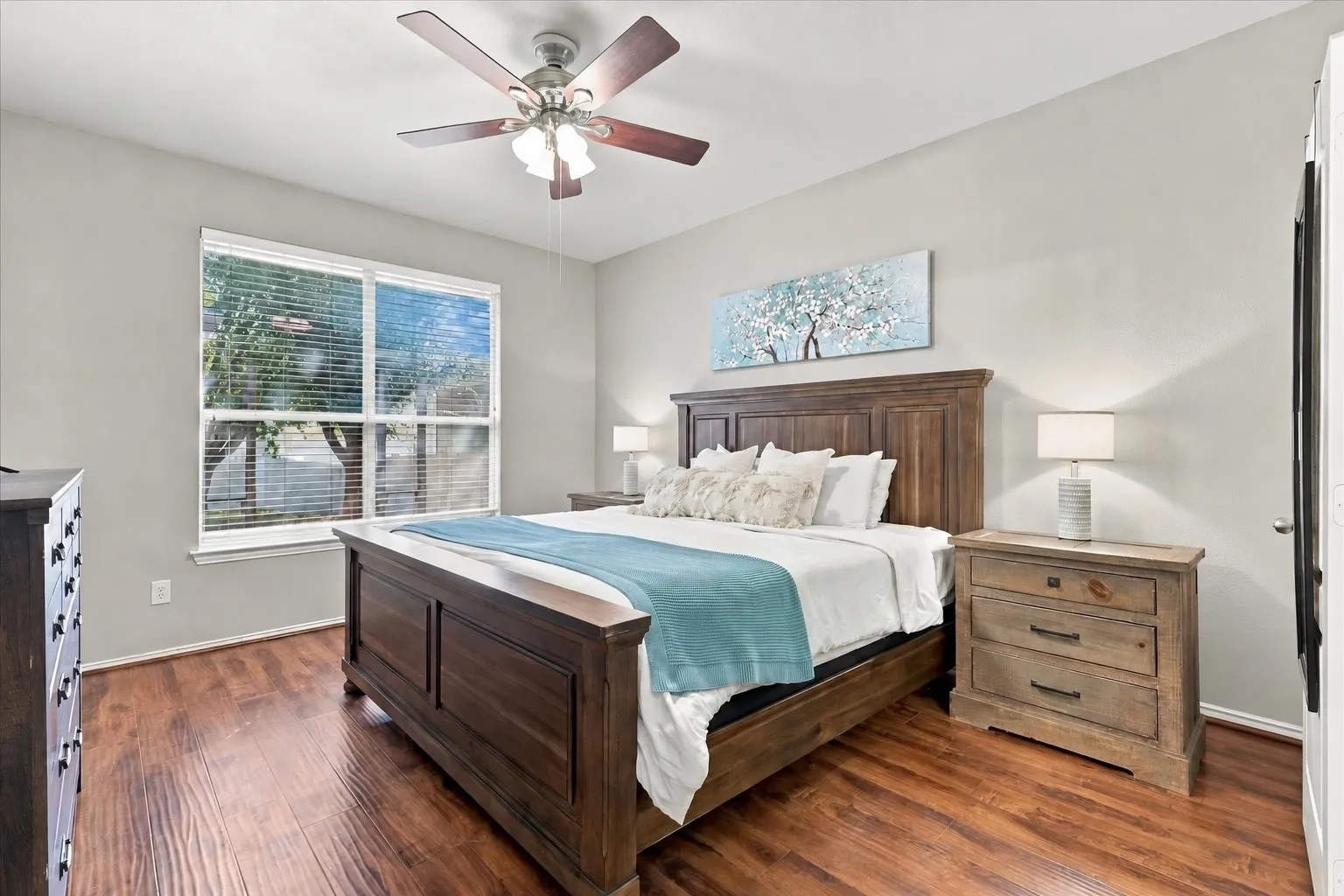 Wood-finish flooring, a large window with blinds, a ceiling fan with integrated lighting, and a light-toned wall finish