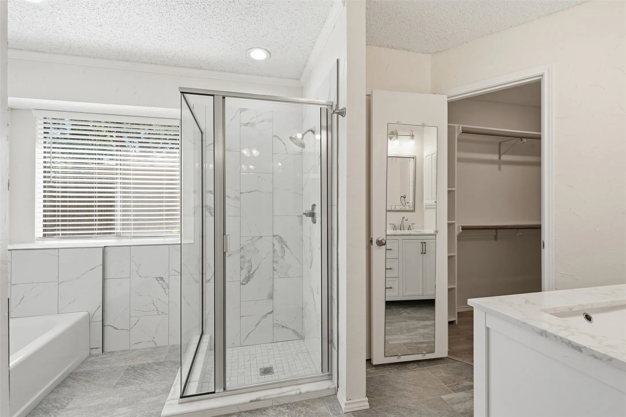 Walk-in shower with clear glass enclosure and subway tile surround, white bathtub, window with horizontal blinds, gray tile flooring, and a vanity with a white countertop