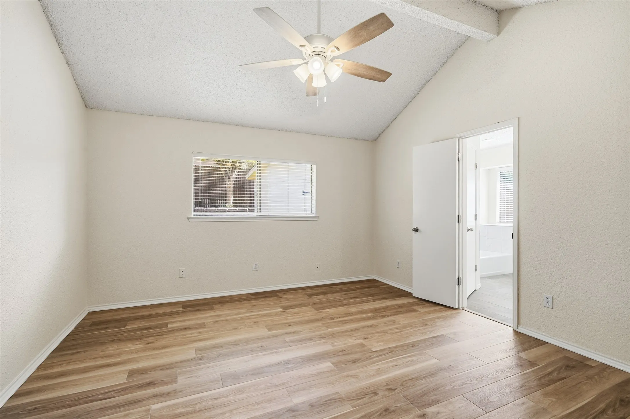 Vaulted ceiling with exposed beam, ceiling fan with light fixture, wood-finish flooring, white horizontal blinds, and a white interior door