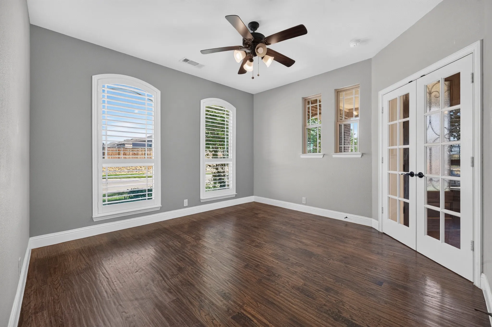 Bright corner room featuring gleaming wood-finish flooring, ceiling fan with integrated lighting, and expansive windows with plantation shutters flooding the space with natural light