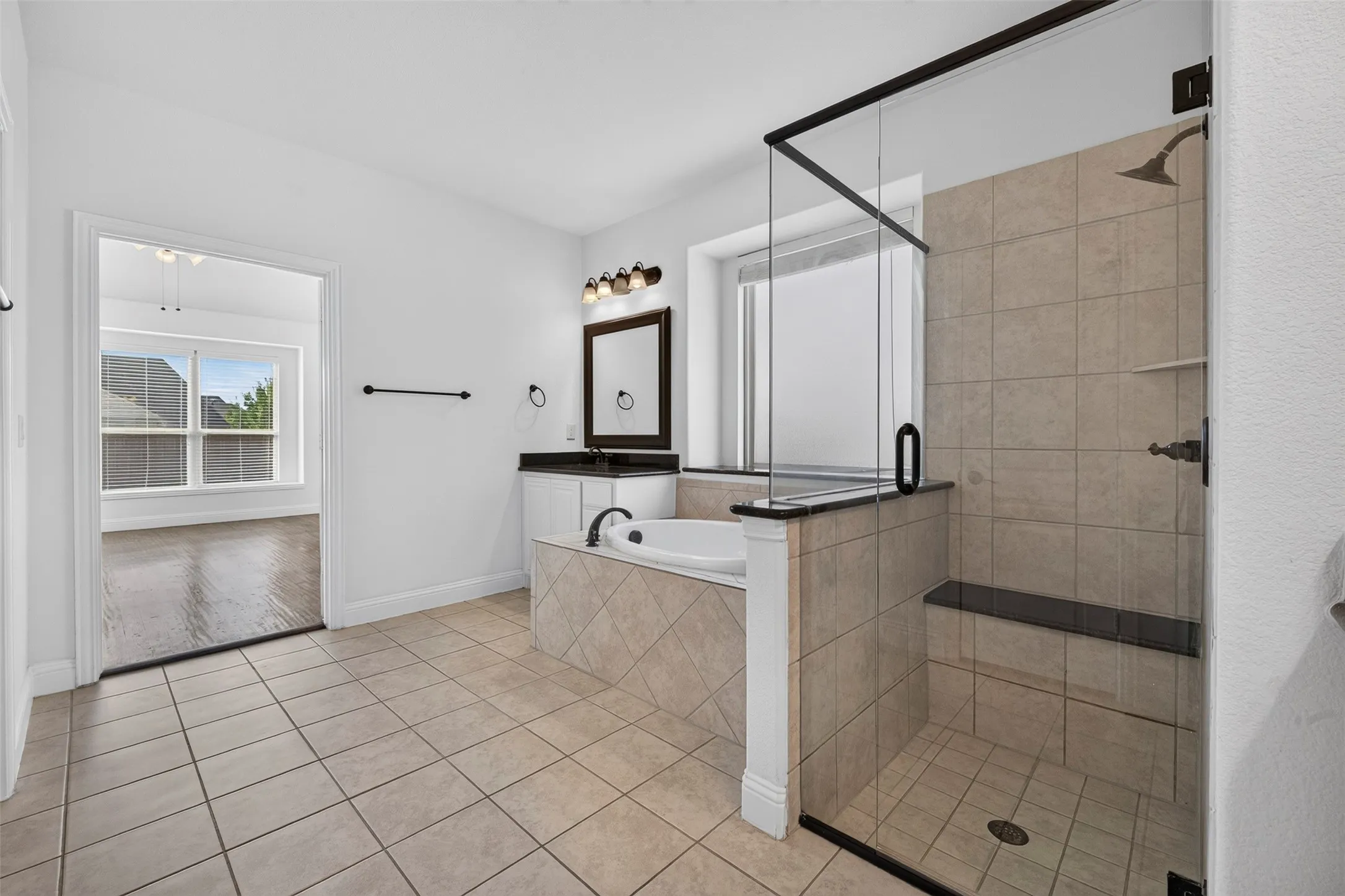 Spacious bathroom featuring tile flooring, a glass-enclosed shower with a built-in bench, a separate soaking tub, and a vanity with a dark countertop