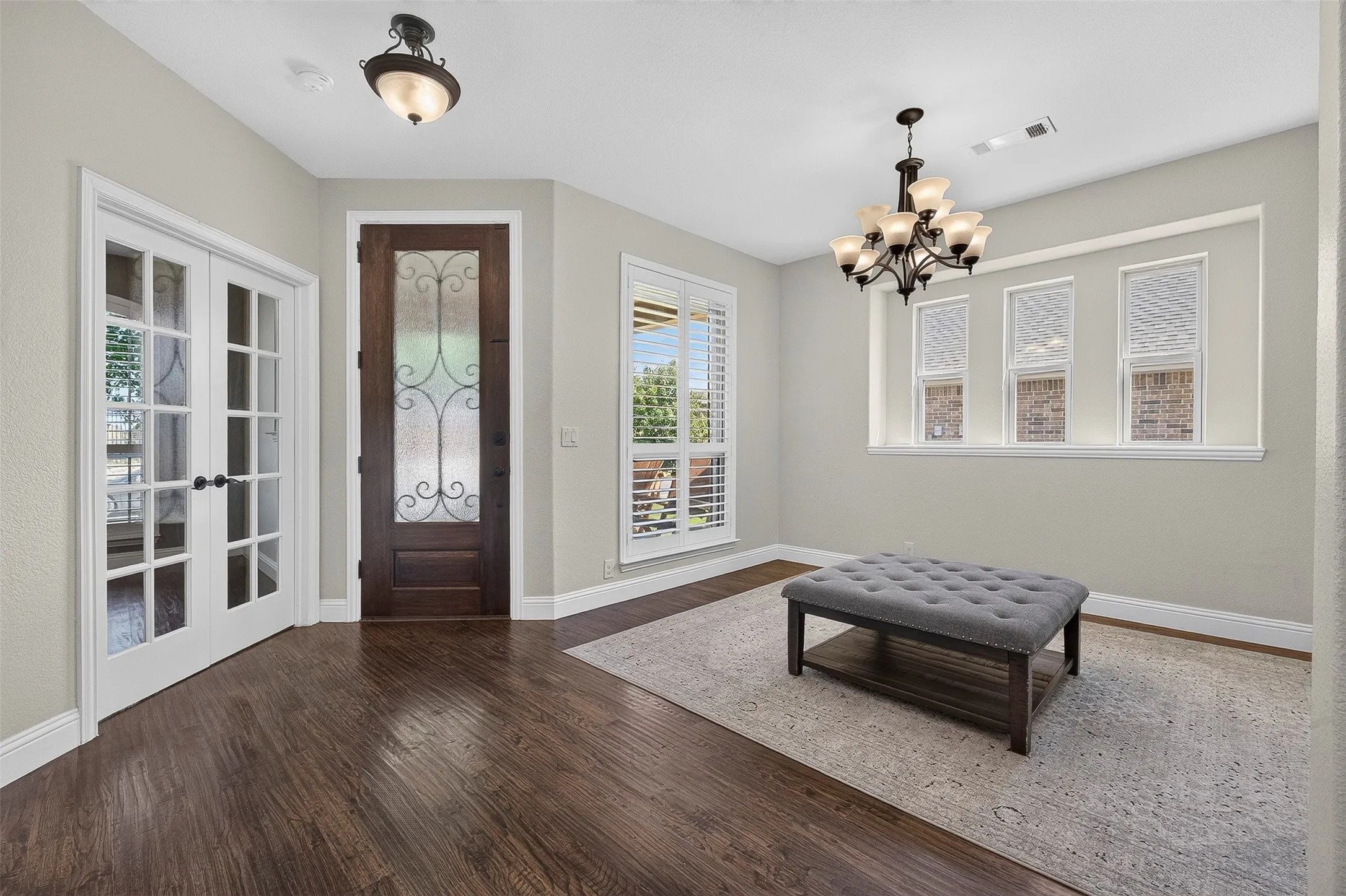 Bright, light-filled foyer showcasing dark wood-finish flooring, white French doors with glass panes, shuttered windows with natural light, a bronze chandelier, and recessed ceiling lighting