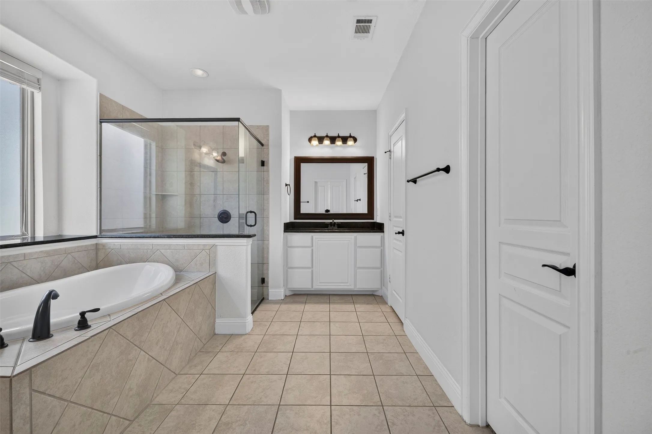 Spacious bathroom featuring a built-in soaking tub, glass-enclosed shower, single vanity with a dark countertop, and tiled flooring