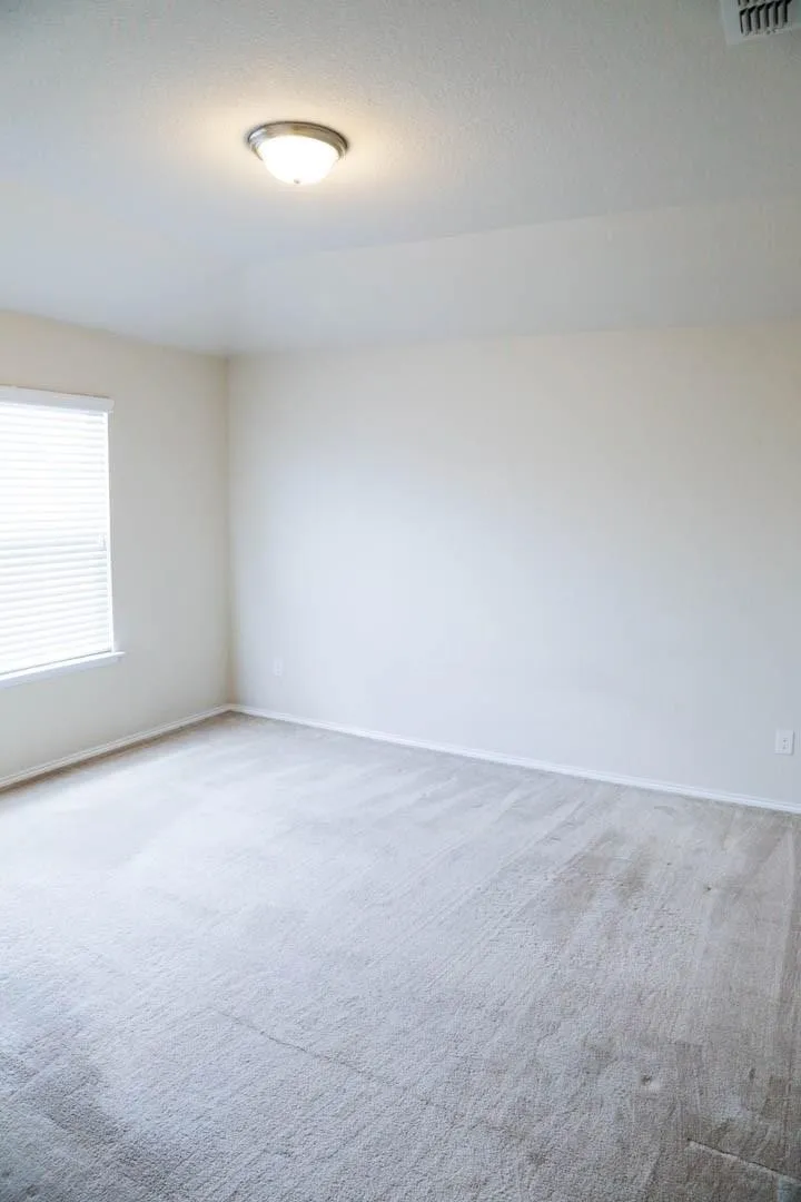 White walls with light-toned carpet, featuring a window with horizontal blinds and a flush-mount ceiling light