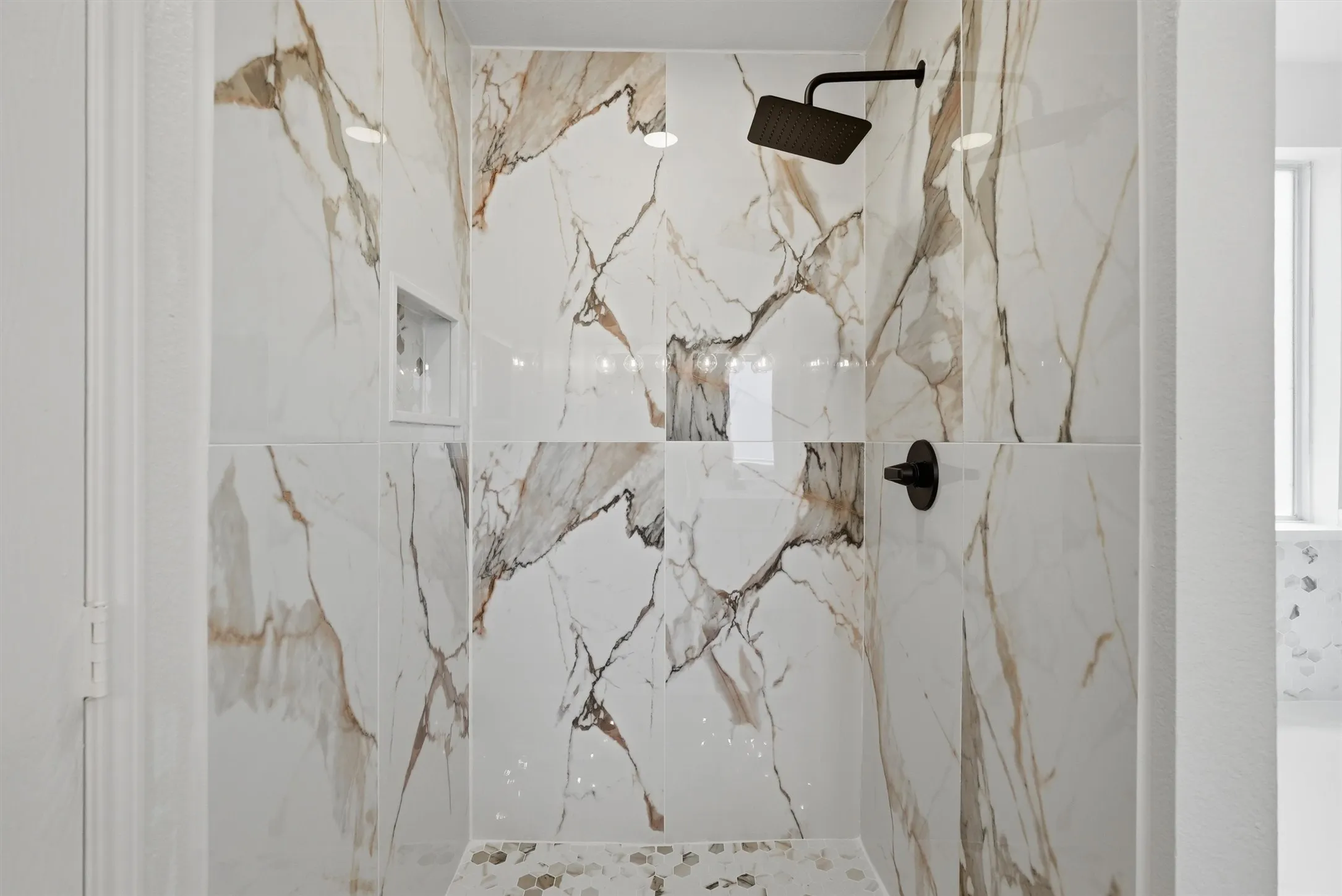 Shower with large format marble-look tile, matte black shower fixtures, a built-in shower niche, and a mosaic tile floor