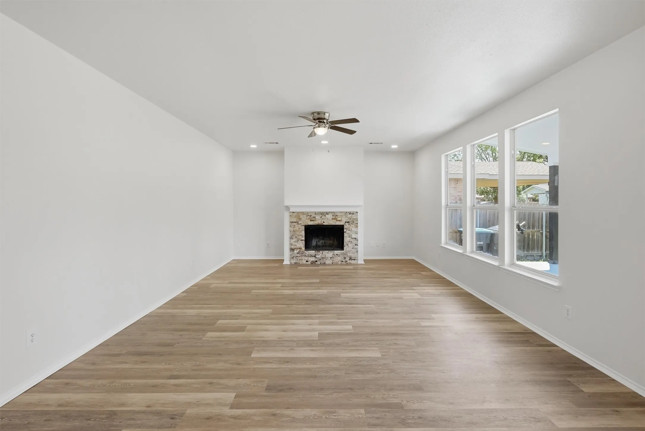 Spacious room featuring wood-finish flooring, a stone-faced fireplace with a white mantel, and multiple windows
