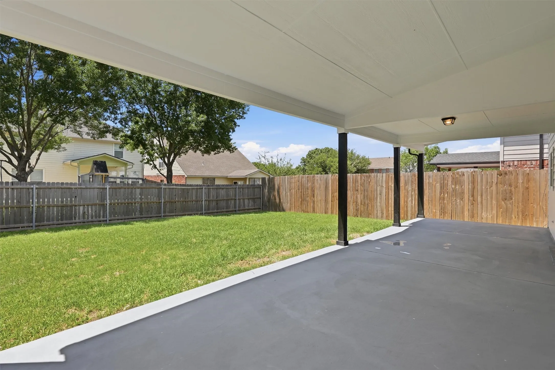 Expansive covered patio with a concrete surface, supported by dark-toned posts, overlooking a fenced yard with a manicured lawn