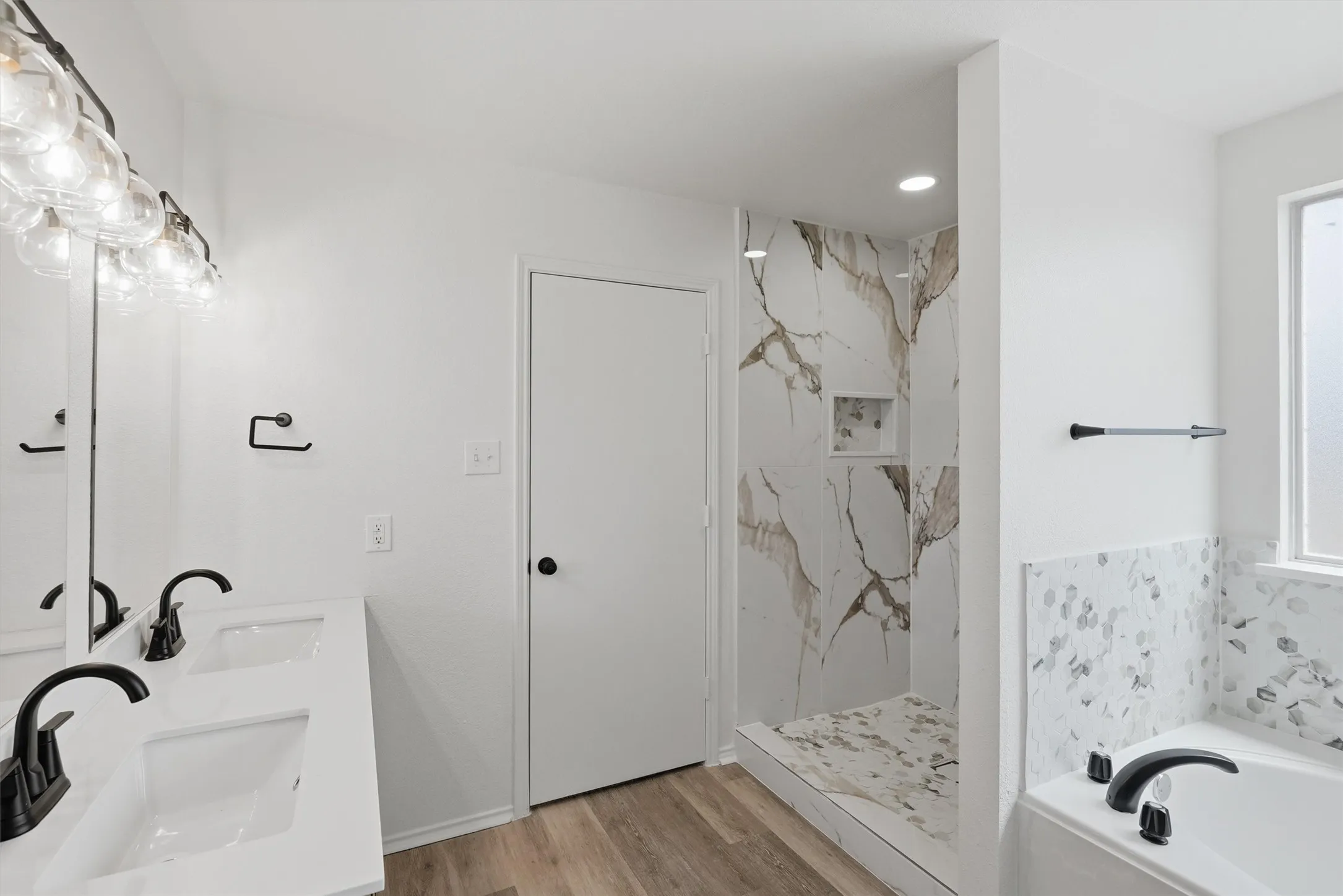 Dual vanity with matte black faucets, wood-finish flooring, walk-in shower with integrated niche, and built-in tub with tile surround