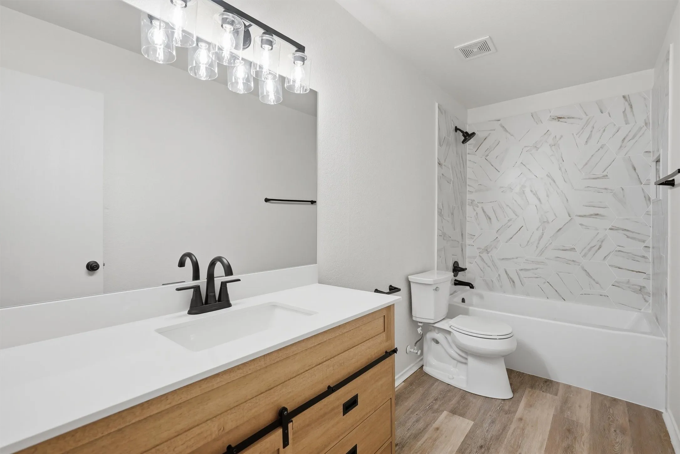 Wood-finish flooring, white vanity countertop, wood-look vanity with black hardware, white tub with patterned surround, and matte black fixtures