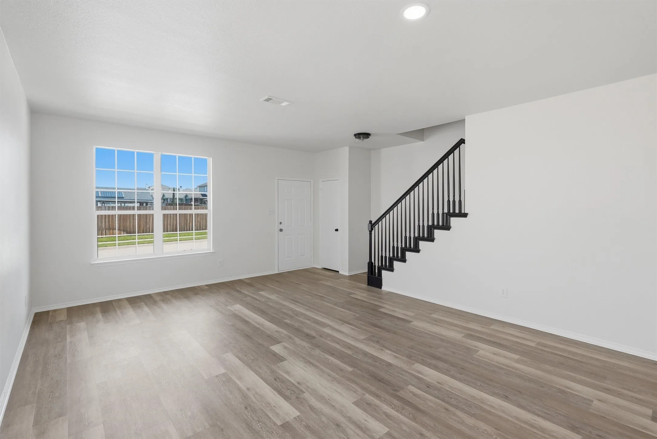 Spacious living area featuring wood-finish flooring, a large window with grid pattern, white walls, and a staircase with black railings