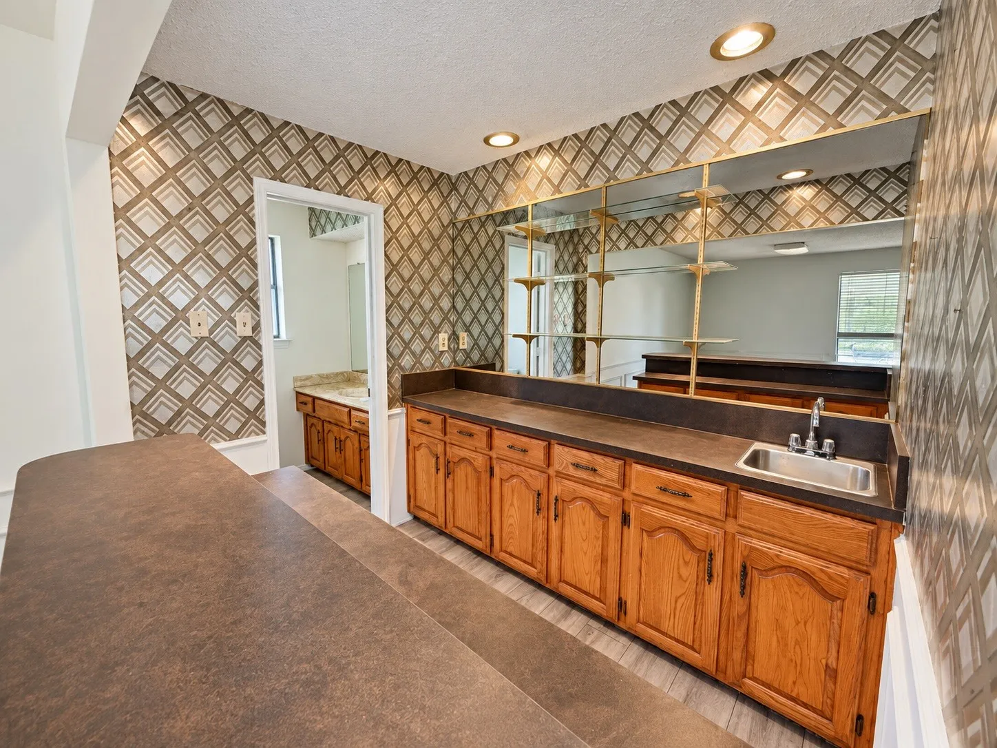 Built-in cabinetry with a stainless steel sink, large mirrored wall with shelving, recessed lighting, and patterned wall covering