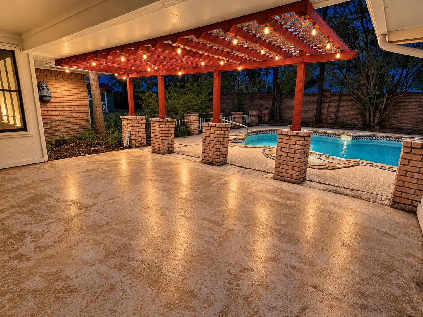 Covered patio featuring a red pergola with string lighting, brick support columns, textured concrete flooring, and a swimming pool with stone coping
