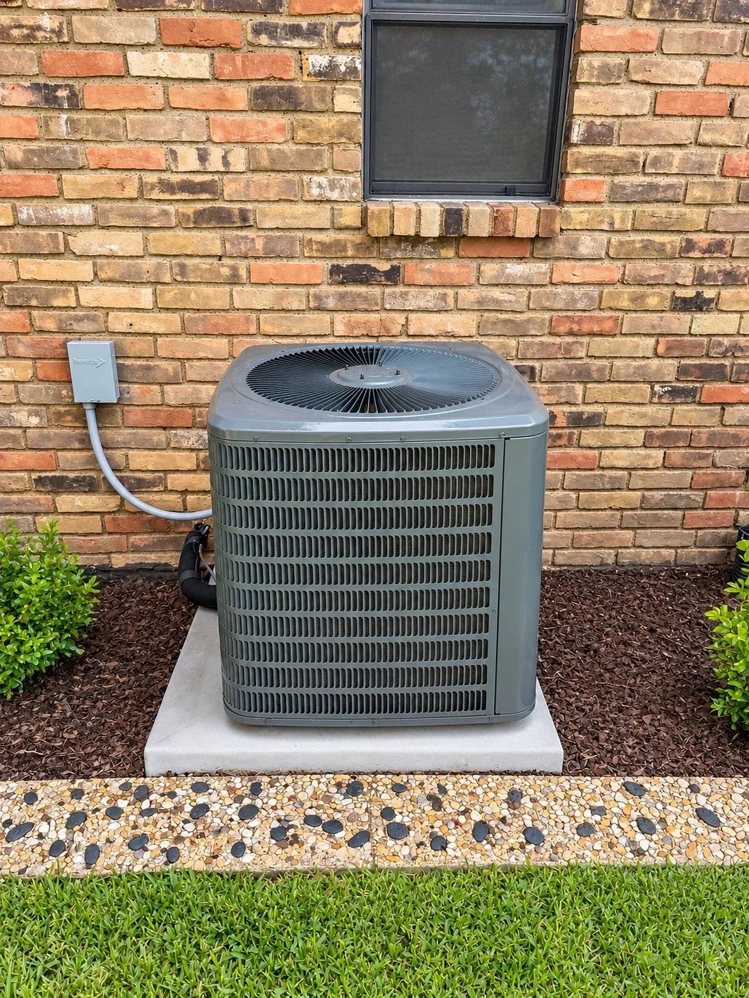 Brick exterior with a window, air conditioning unit on a concrete pad, decorative stone border, and landscaping