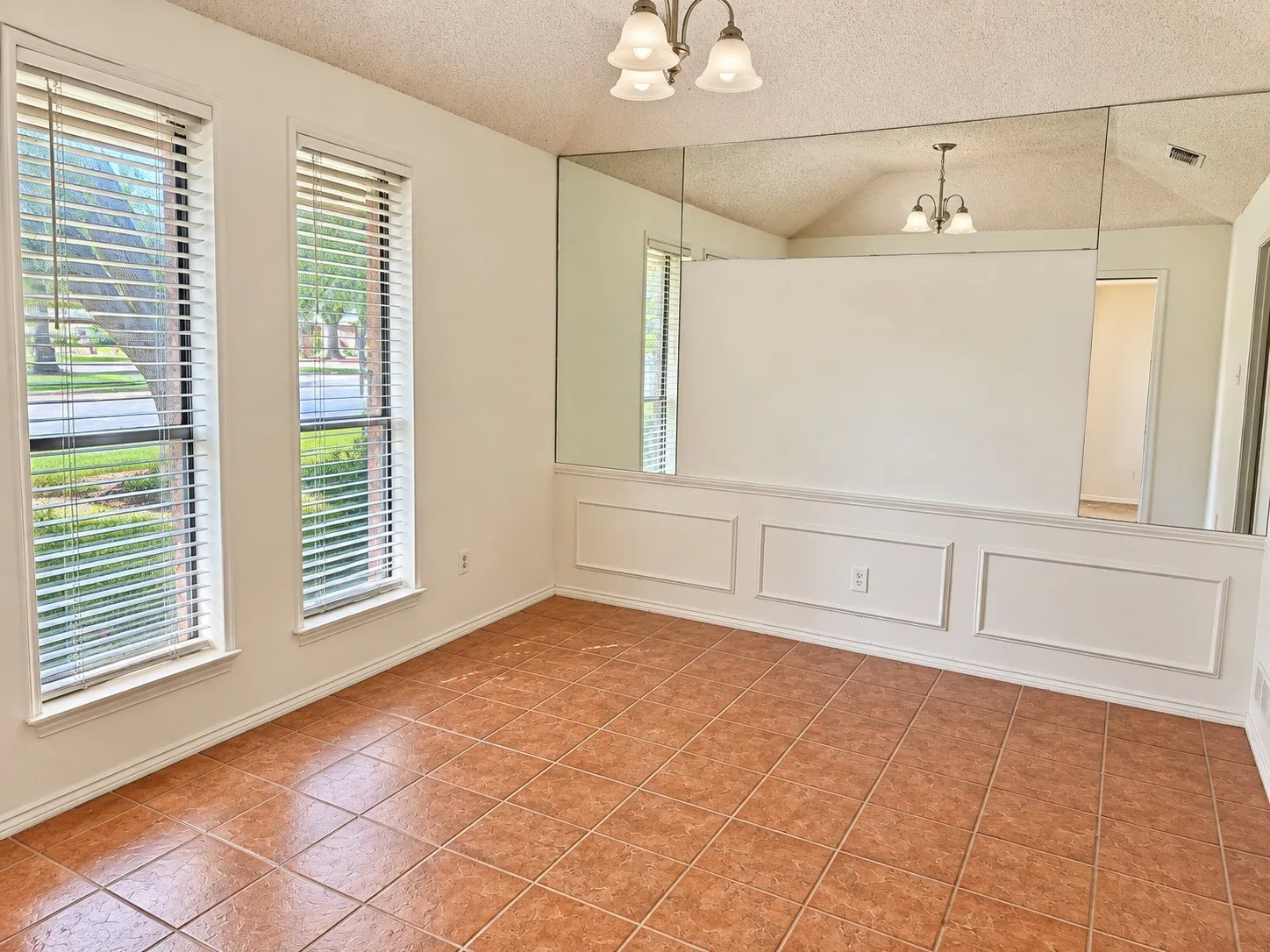 Terracotta-style tile flooring, two window openings with blinds, decorative wall molding, full-height wall mirror, and a ceiling-mounted light fixture