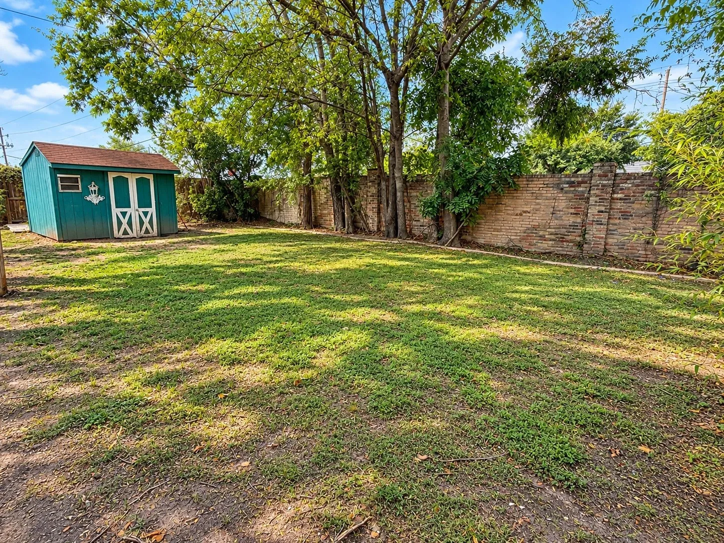 Expansive grassy yard with mature trees, a brick perimeter wall, and a separate storage shed with double doors