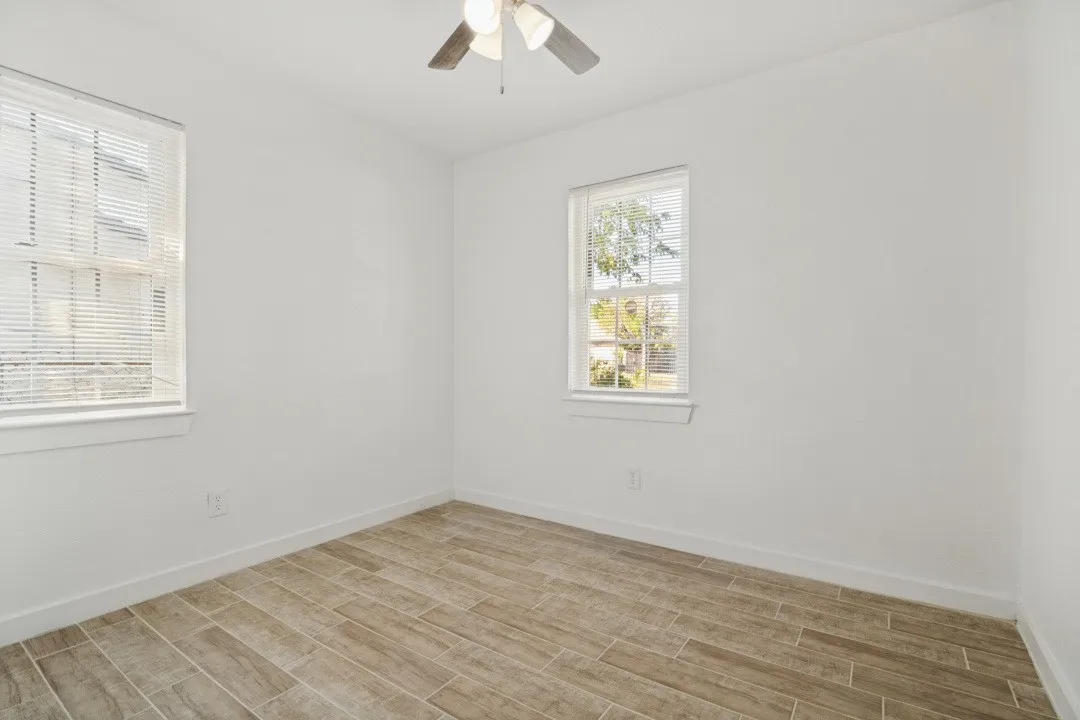 Wood-finish tile flooring, two windows with blinds, ceiling fan with integrated lighting, and white walls