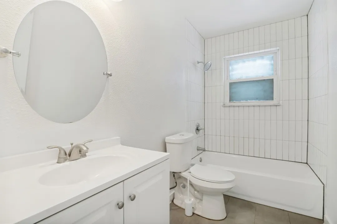 Vanity with integrated sink and brushed nickel faucet, wall-mounted oval mirror, tiled tub-shower combination, and window with privacy glass
