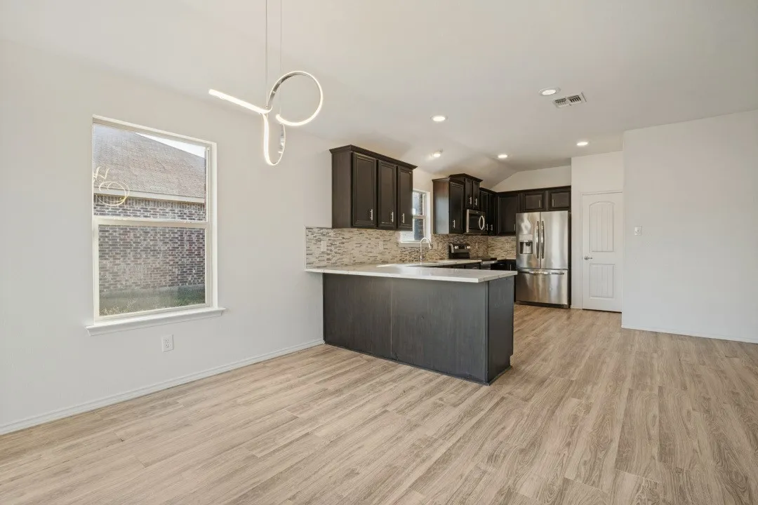 Open-concept living space featuring wood-finish flooring, a contemporary pendant light fixture, dark wood cabinetry, a mosaic tile backsplash, and stainless steel appliances