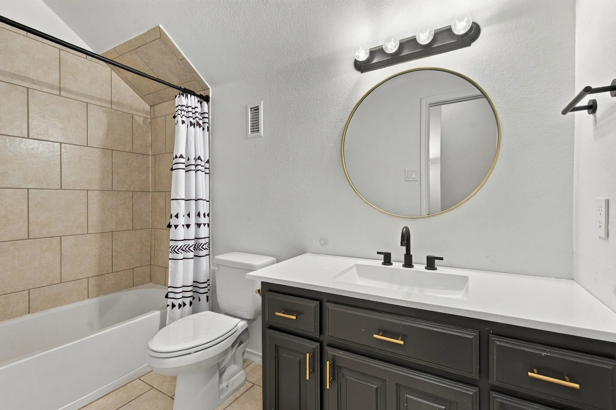 Bathroom featuring a tub with tile surround, a modern vanity with a white countertop and matte black fixtures, and a round wall mirror