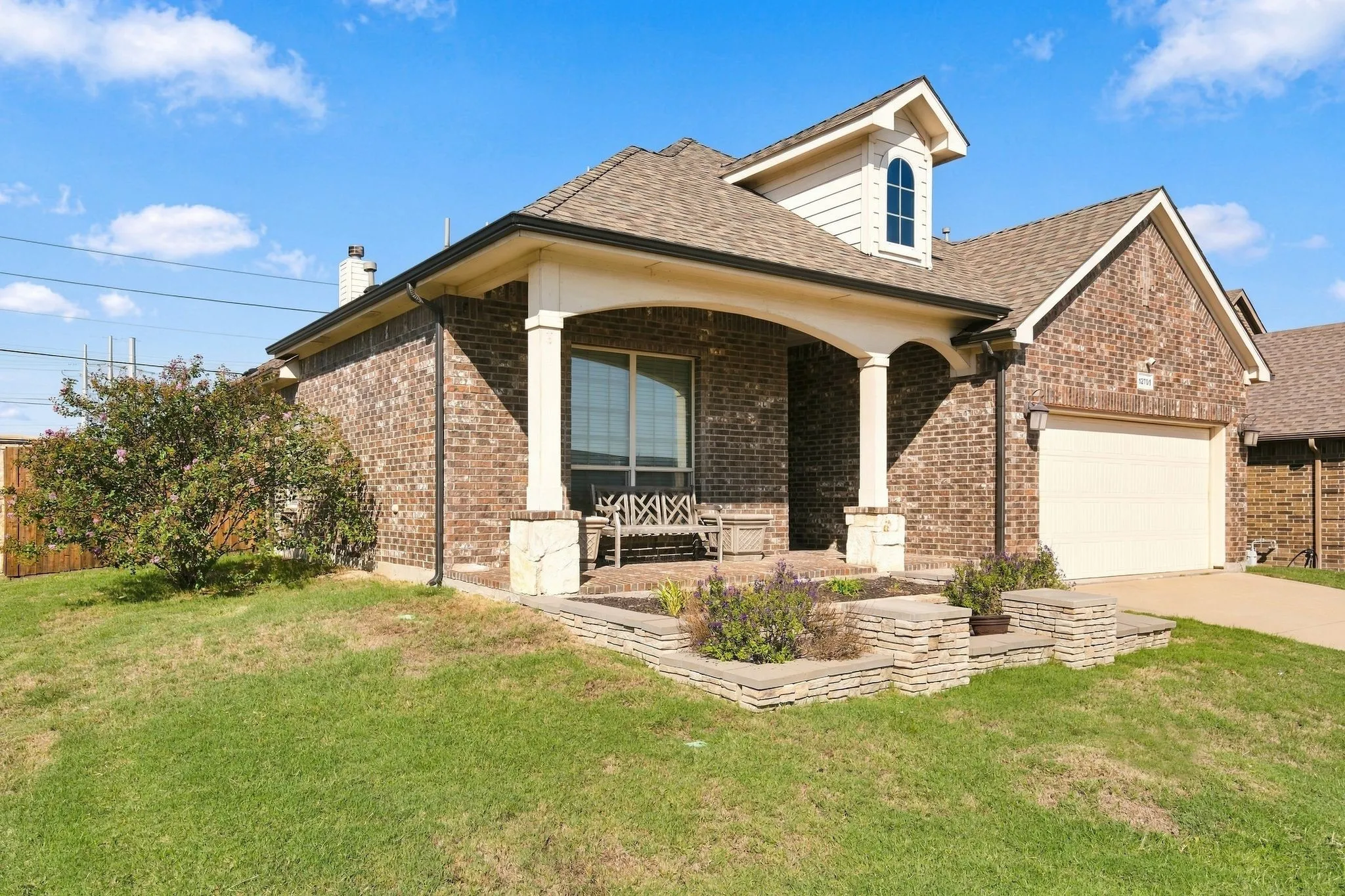 Brick exterior with a covered porch, dormer window, attached garage, and stone-clad landscaping