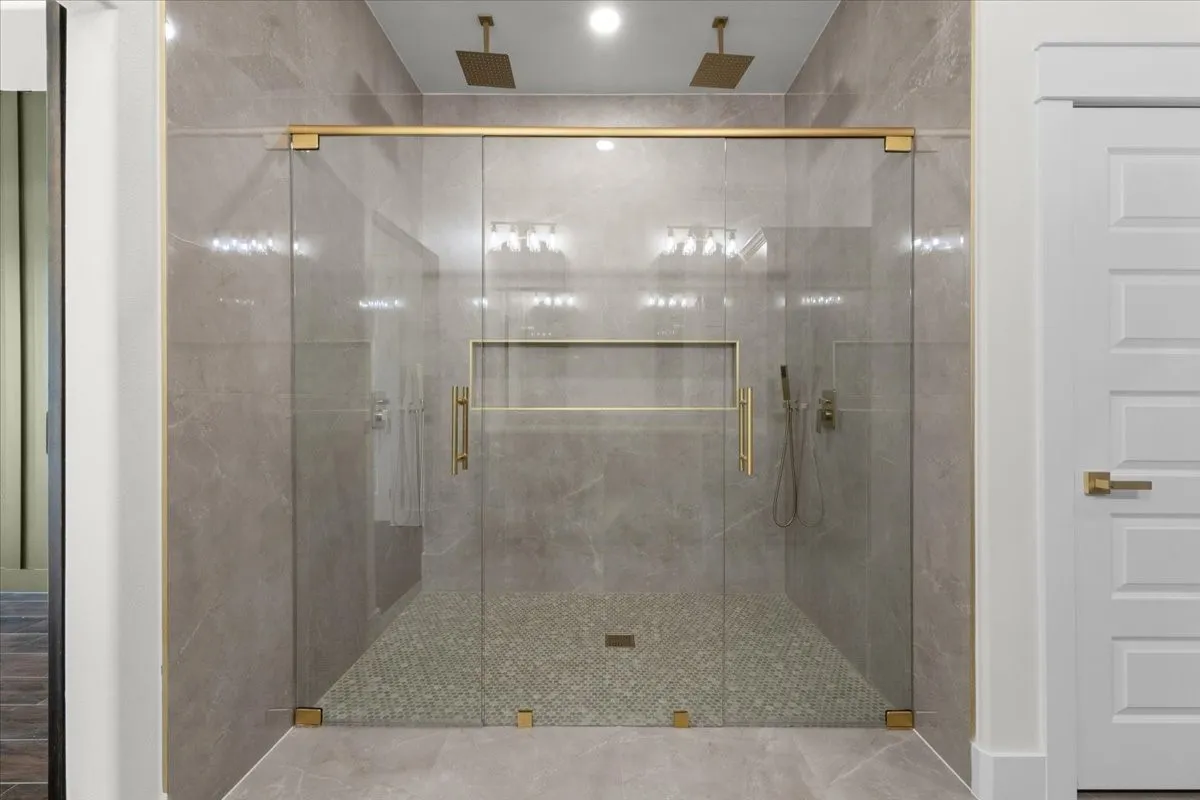 Frameless glass shower enclosure with brass-finish hardware, dual ceiling-mounted rain showerheads, a recessed shower niche, and full-height stone-look tile