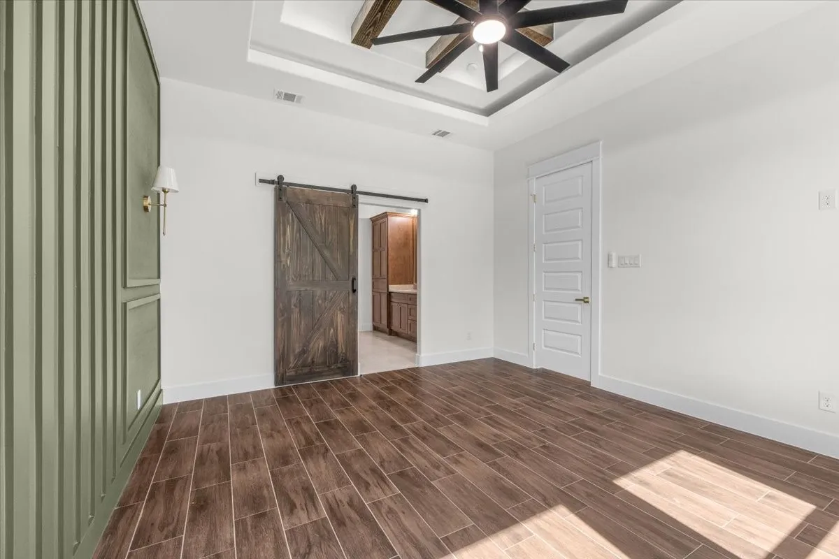 Tray ceiling with a dark-tone wood-finish ceiling fan, wood-finish flooring, a contemporary white interior door, a dark wood-finish barn door, and a paneled accent wall with a sconce