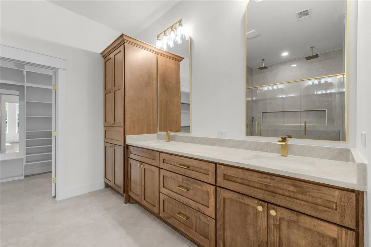 Dual vanity with light-toned countertops, wood-finish cabinetry, and gold-finish fixtures