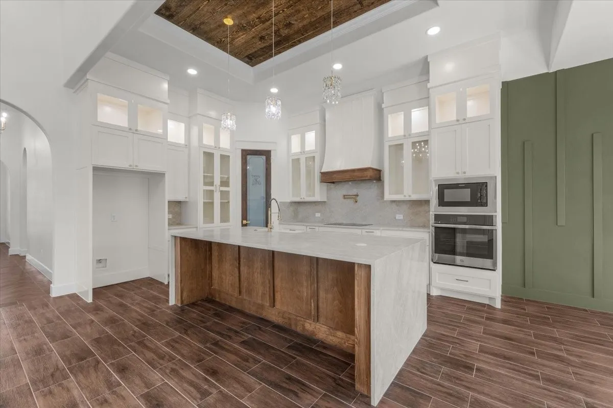 Kitchen featuring wood-finish flooring, a central island with a waterfall countertop, white cabinetry with glass-front uppers, a built-in oven and microwave, and a wood-paneled ceiling