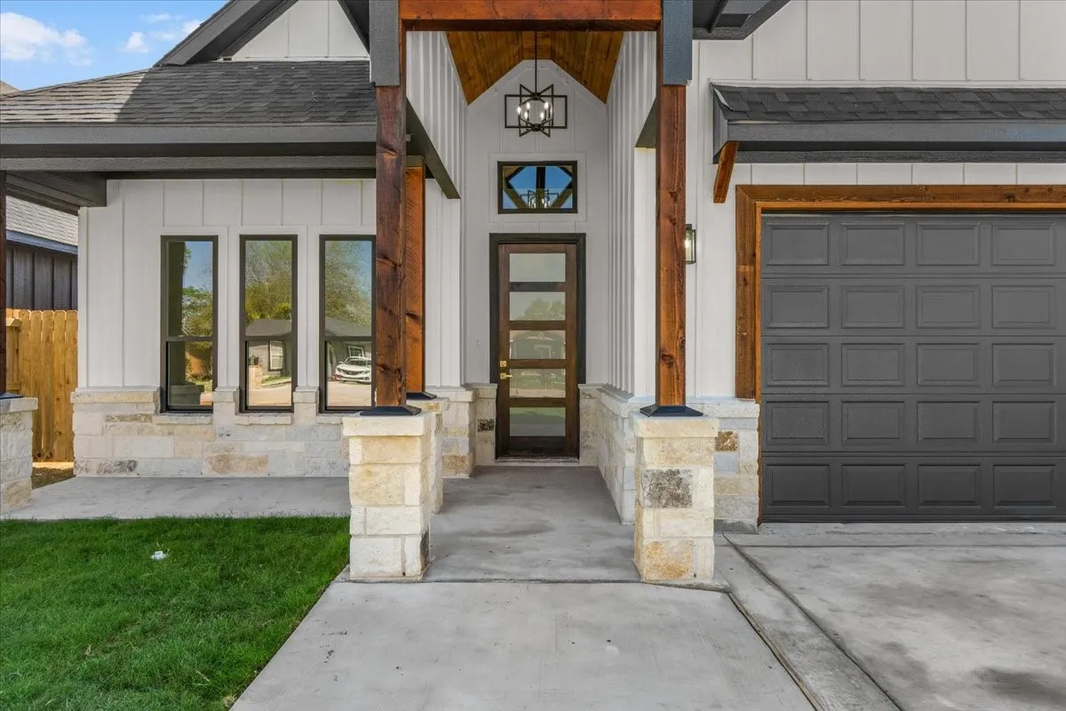 Modern farmhouse exterior with white vertical siding, natural stone accents, dark-framed windows, a wood-paneled garage door, and exposed wood beams