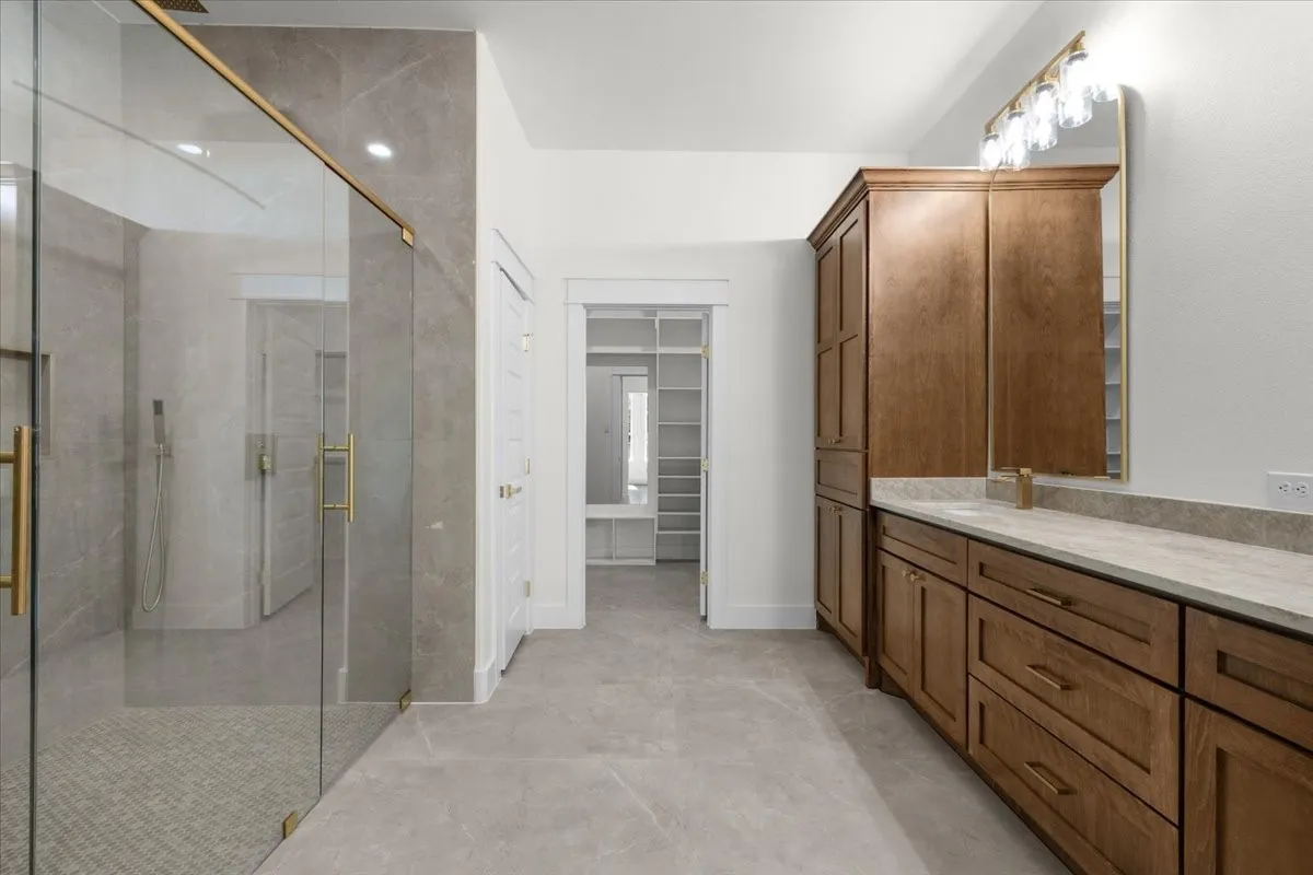 Glass-enclosed shower with gold-tone hardware, a long wood-finish vanity with ample storage, a framed mirror, a five-bulb vanity light, and recessed ceiling lighting