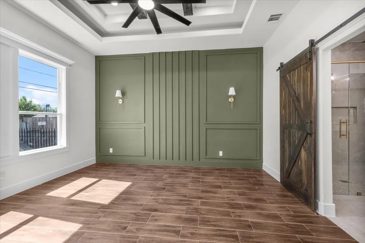 Tray ceiling with a ceiling fan, accent wall with sconce lighting, wood-finish flooring, and a sliding barn door