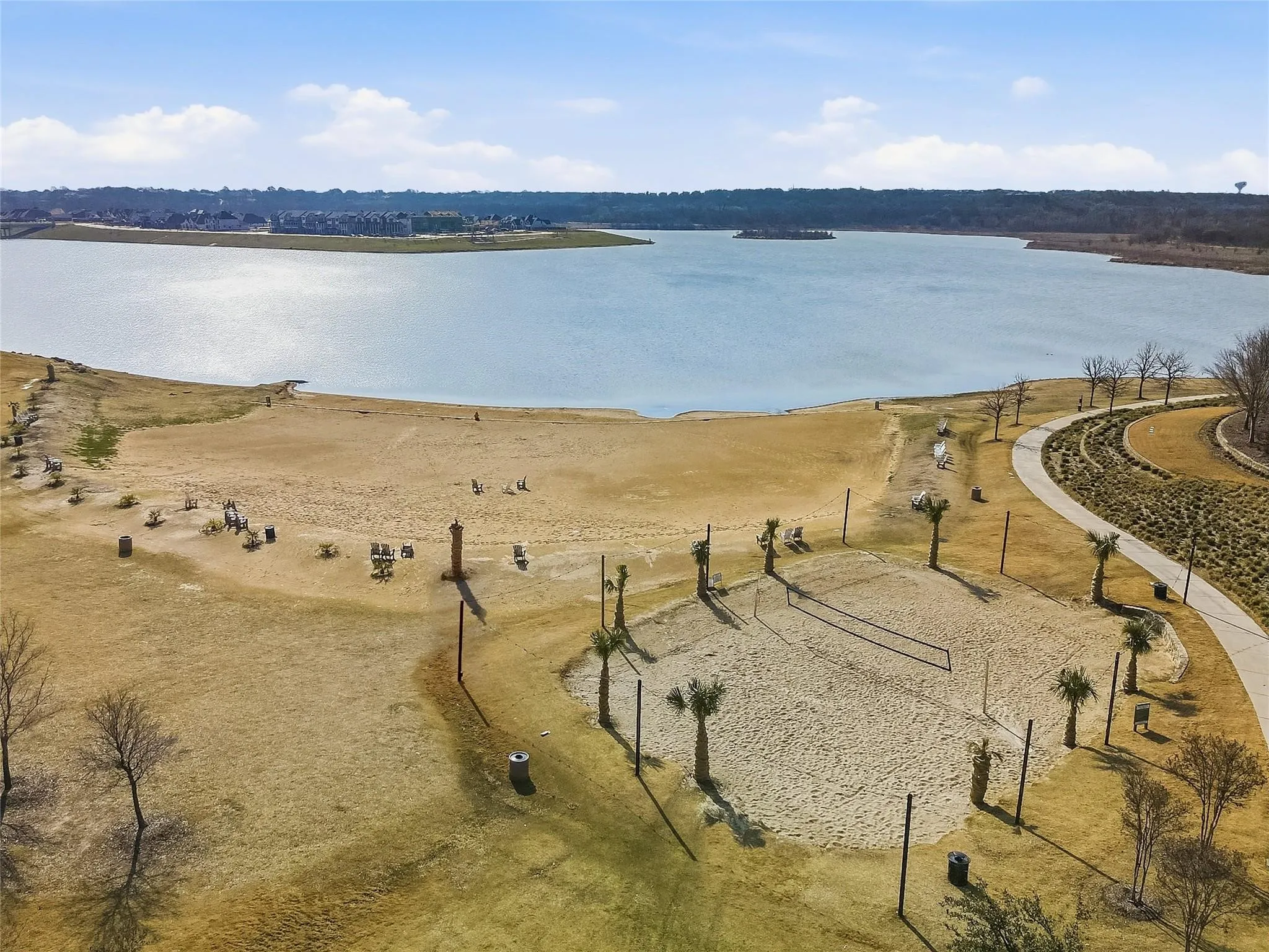 Lakefront property featuring a sandy beach area, dedicated sand volleyball court, established palm trees, and an extensive paved pathway