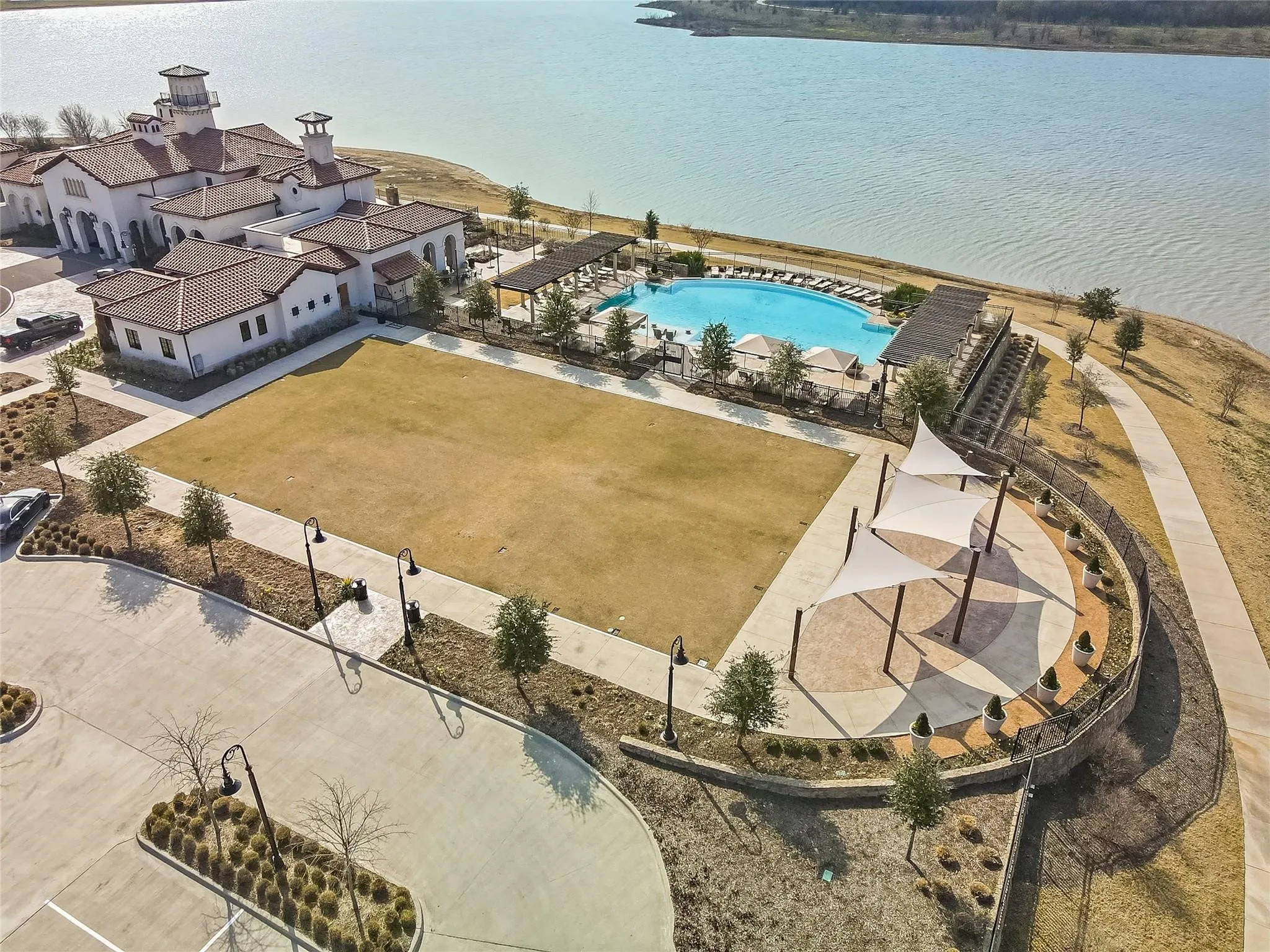 Waterfront property featuring a large resort-style pool, expansive green space, shaded outdoor seating, and a multi-level clubhouse with a tile roof