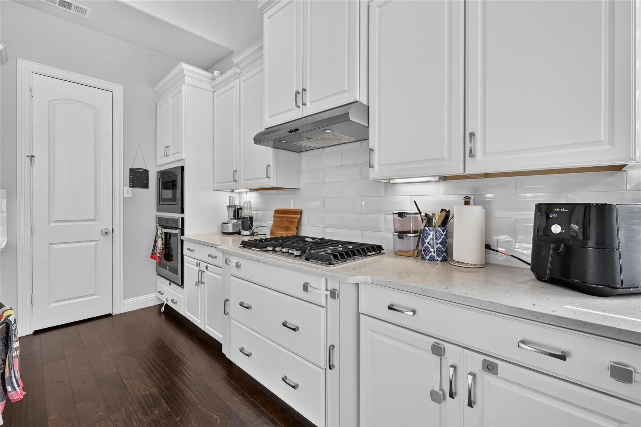 White shaker-style cabinetry, subway tile backsplash, gas cooktop, built-in double ovens, and wood-finish flooring