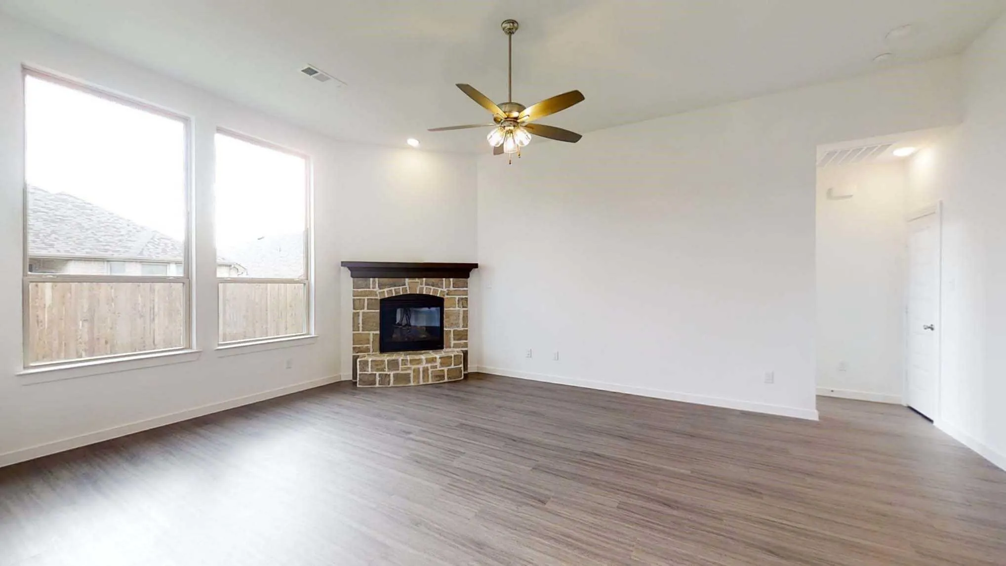 Spacious room featuring wood-finish flooring, a corner fireplace with a stone surround and wood mantel, multiple windows, and a ceiling fan with integrated lighting