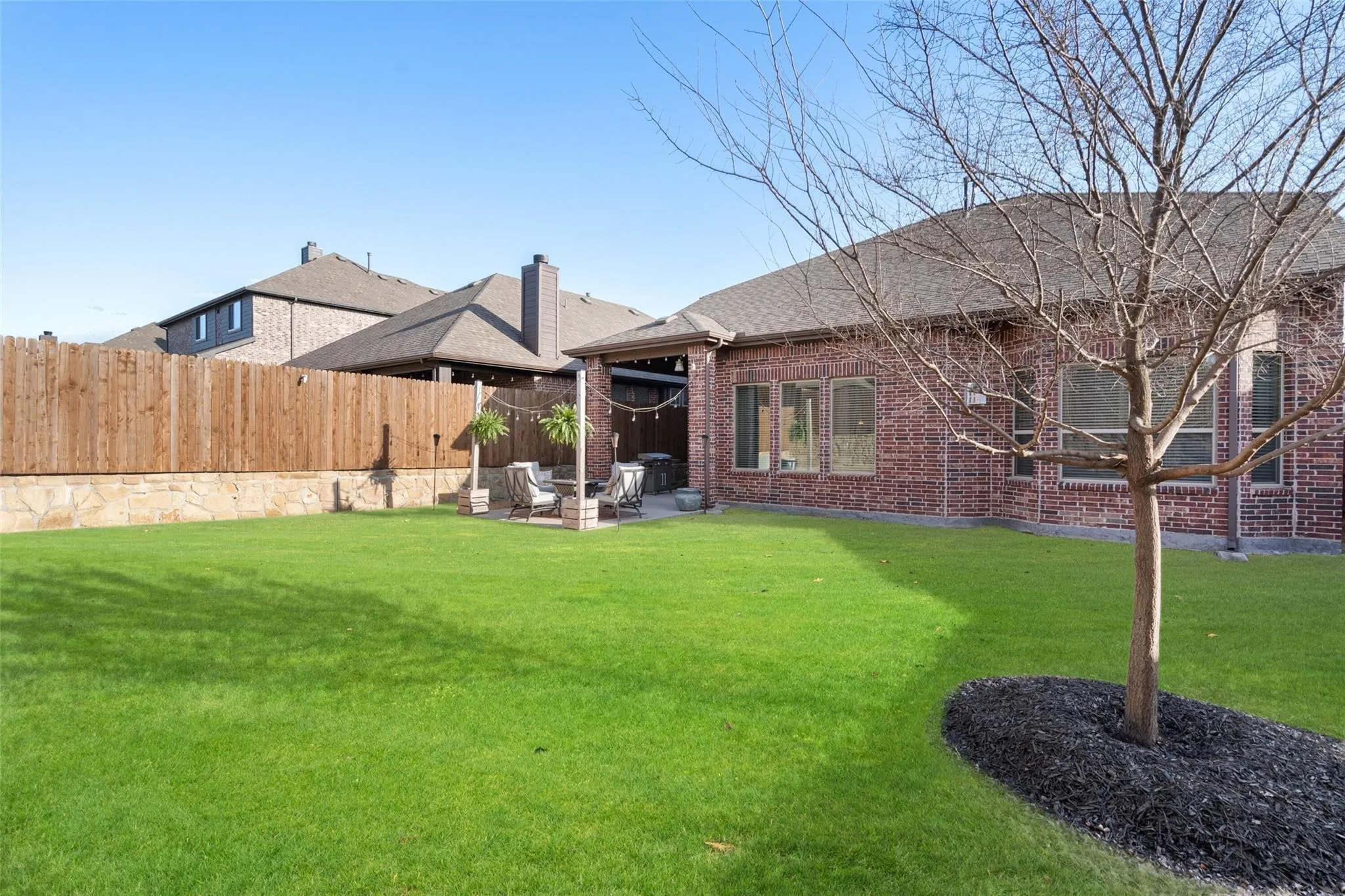 Expansive backyard featuring a manicured lawn, brick exterior, covered patio, and privacy fencing with stone foundation
