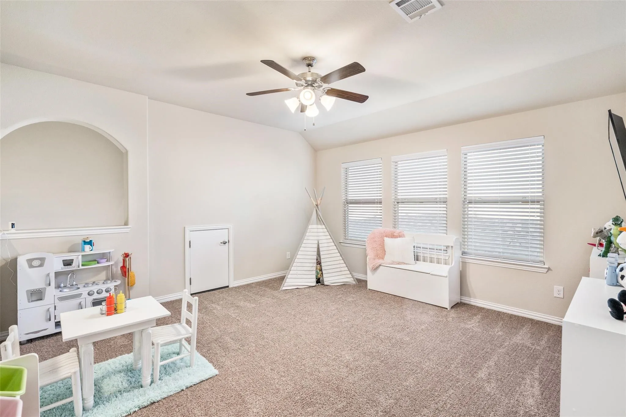 Carpeted room with a ceiling fan, three windows with blinds, a white door, and an arched wall opening